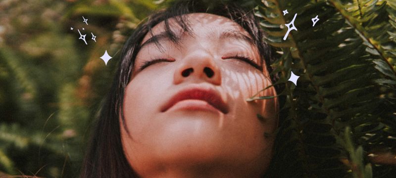 Woman with black hair with ferns over her face smiles softly with her eyes closed